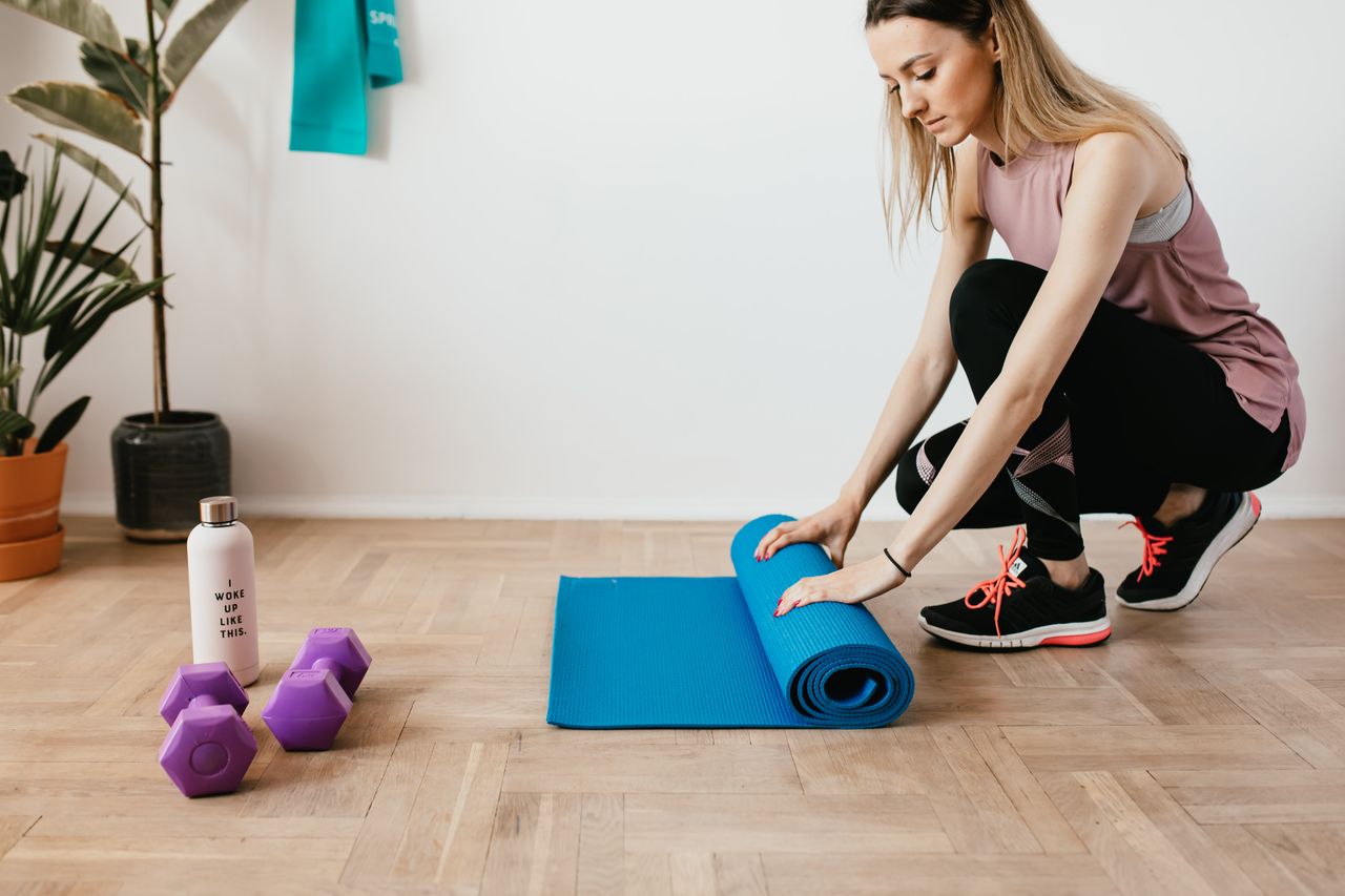 Side view of young female athlete in sportswear unfolding blue sport mat and looking down while sitting on floor near dumbbells and water bottle preparing for training indoors