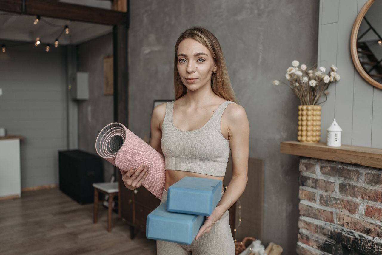 A woman in a sports bra holds yoga mat and blocks, standing in a modern studio setting.